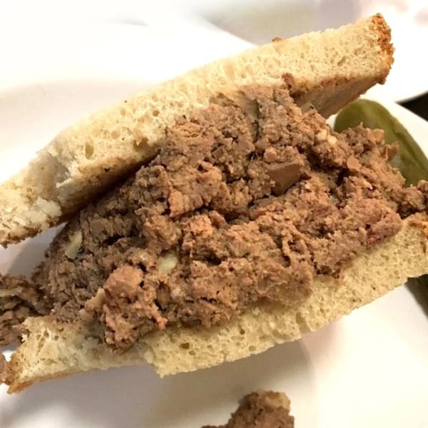 A Helfeld's Delicatessen chopped liver sandwich on white bread, with a pickle spear in the background.