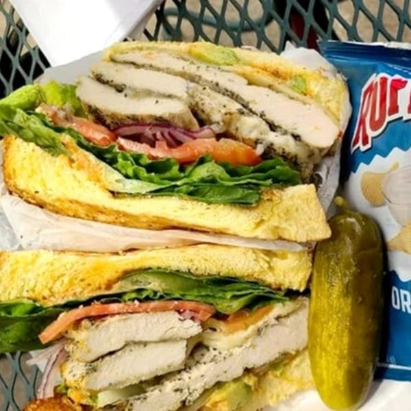Helfeld's Delicatessen chicken sandwich with lettuce, tomato, and onion, served with a pickle and chips.