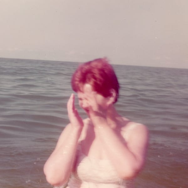A blurry, vintage photo of a woman with red hair in the ocean, covering her face.