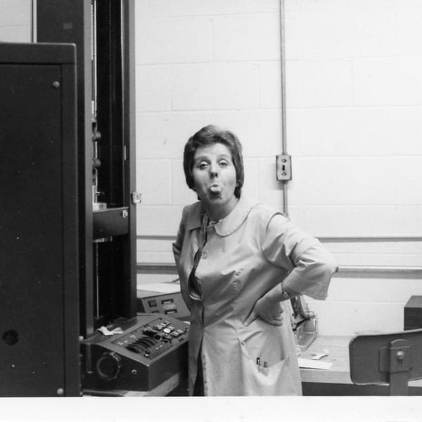 A woman in a light-colored coat sticks her tongue out while standing in a lab.