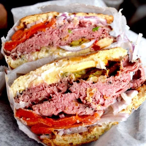 A Helfeld's Delicatessen sandwich, cut in half, revealing layers of pastrami, tomatoes, and onions.